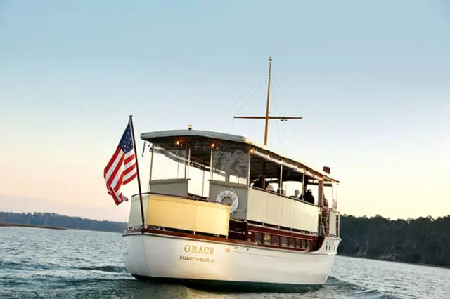 The yacht 'Grace' sails on calm water with an American flag flying from the stern. The boat has a white hull, mahogany trim, and an open upper deck with seating.