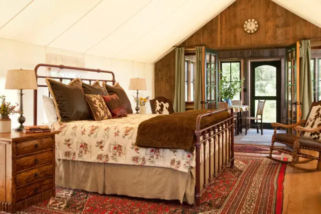 Bedroom with a metal bed, patterned comforter, brown pillows, and a rustic wooden nightstand. A rocking chair and a small table with chairs are visible near French doors leading outside.