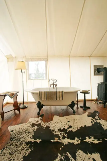 Tent interior with a clawfoot bathtub, wood floor, cowhide rug, vintage floor lamp, and a black wood-burning stove.