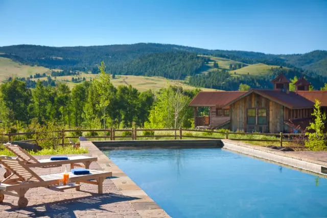 Infinity pool with lounge chairs and drinks overlooking a rustic wooden building and rolling green hills under a clear blue sky.