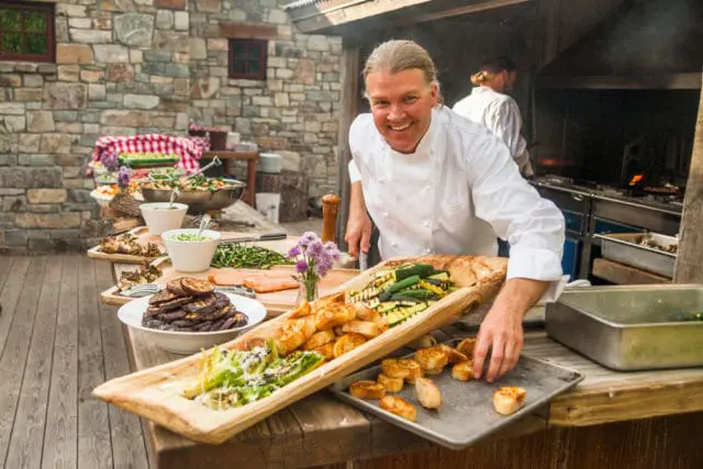 Chef in white uniform smiles while preparing a buffet of grilled vegetables, salmon, and bread outdoors with a stone building in the background.