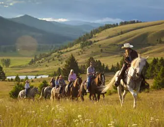 Group of people on horseback riding through a grassy field with rolling hills and a small lake in the background under a hazy sky