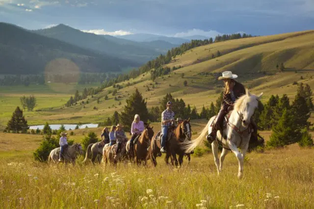 Group of people on horseback riding through a grassy field with rolling hills and trees in the background under a cloudy sky.