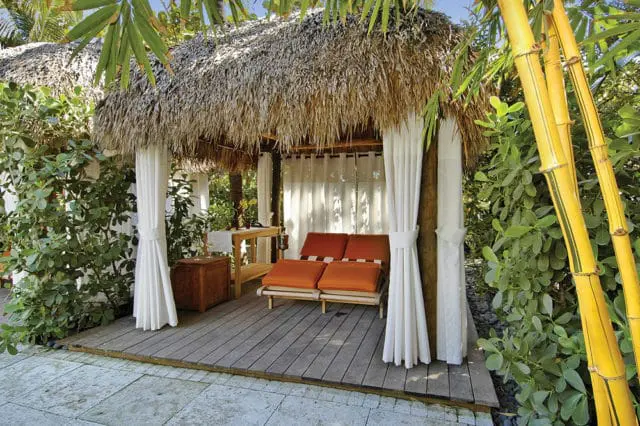 Tiki cabana with thatched roof, white curtains, and orange lounge chairs on a wooden deck, surrounded by lush green foliage and bamboo stalks.