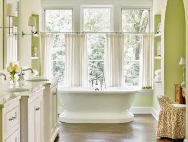 Light green bathroom with a white freestanding bathtub, double vanity with marble countertops, and a patterned chair next to a wooden dresser.