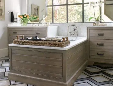 Bathroom with a freestanding tub, wood-paneled vanity, marble countertops, and geometric floor tiles. A large window overlooks trees.