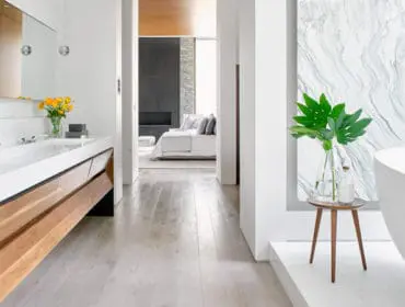 Modern bathroom with a white countertop double vanity, wood cabinetry, and a freestanding bathtub next to a marble accent wall. A view into a bedroom with a fireplace is visible through an archway.
