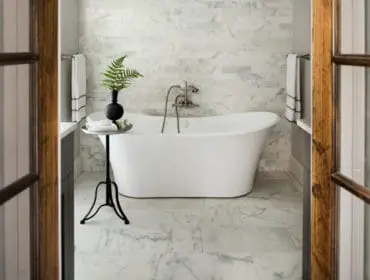 Freestanding white bathtub with marble tile walls and floor, next to a small black side table with a fern and a black vase.