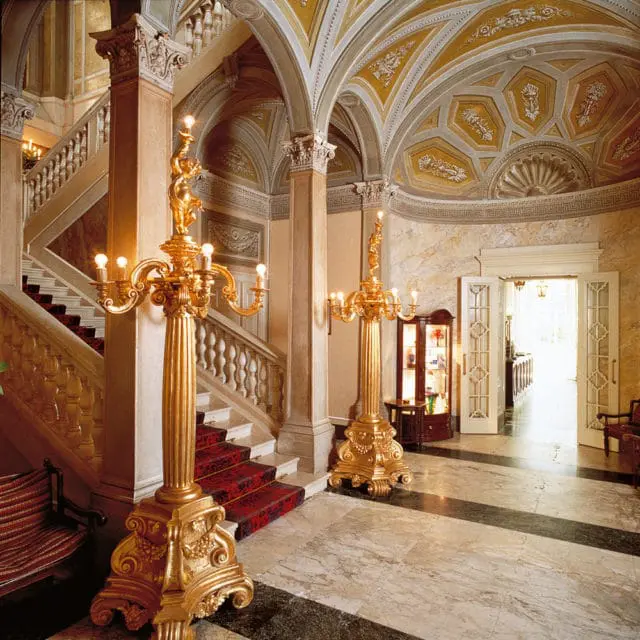 Grand foyer with marble floors, a red carpeted staircase, ornate gold candelabras, and a detailed vaulted ceiling with yellow and white ornamentation.