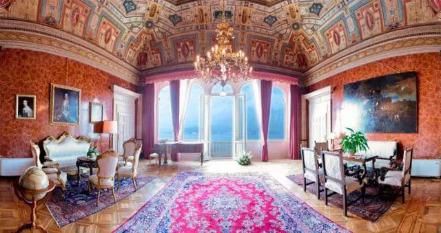 Ornate Italianate living room with red textured walls, gilded furniture, a grand piano, a large crystal chandelier, and panoramic windows overlooking a lake.