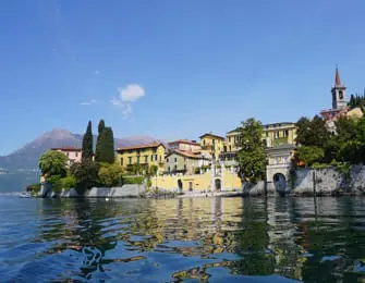 Buildings with yellow facades and red-tiled roofs line a waterfront, with a church steeple visible. Mountains rise in the distance under a clear blue sky.