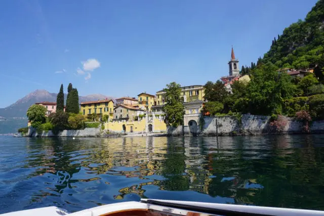 View of a lakeside town with yellow buildings and a church tower, reflected in the water, with a boat in the foreground.