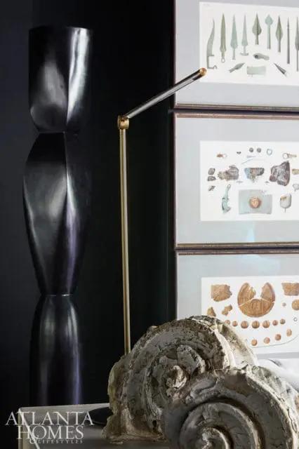 Dark hourglass-shaped sculpture next to a brass floor lamp, with framed illustrations of artifacts and architectural details in the background.