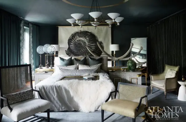 Dark teal bedroom with a large ammonite fossil tapestry above a bed with a fluffy white throw, flanked by cane-backed chairs and a mirrored dresser.