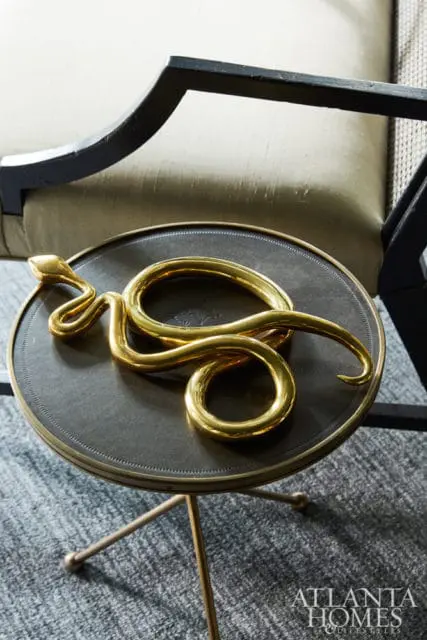 Brass snake sculpture coiled on a round dark metal table with a gold rim, next to a chair with a neutral upholstered seat and black frame.