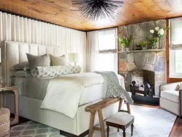 Bedroom with a tufted cream upholstered bed, stone fireplace, wood plank ceiling, and white floor-to-ceiling curtains.