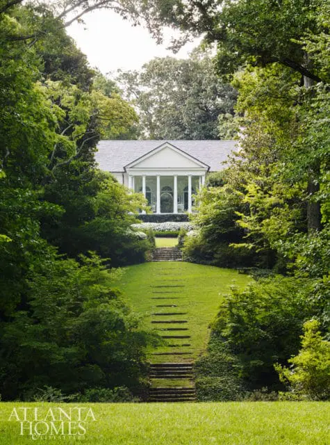 White neoclassical house with a portico supported by columns, surrounded by lush green trees and a terraced lawn with stone steps.