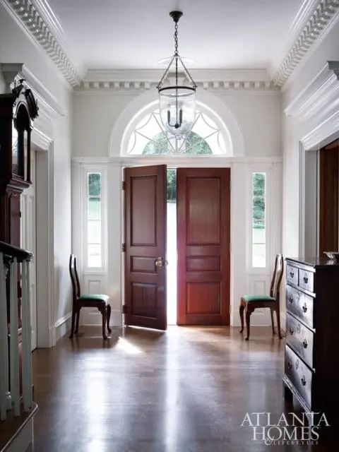 Foyer with double mahogany doors, arched transom window, glass lantern pendant light, and two green upholstered chairs.