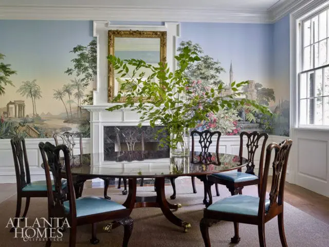 Dining room with oval wood table and upholstered chairs, featuring a scenic mural wallpaper and a fireplace with a gold mirror above.