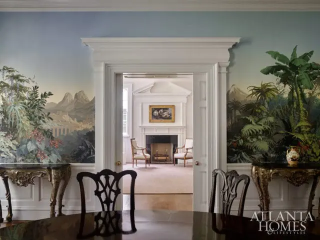 Dining room with ornate wallpaper depicting lush foliage and mountains, leading to a living room with a fireplace and two armchairs.