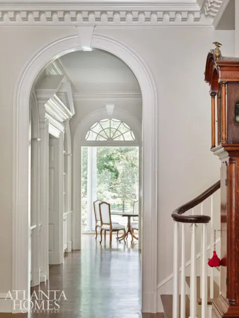 Hallway with arched doorways, white trim, hardwood floors, and a grandfather clock next to a staircase.