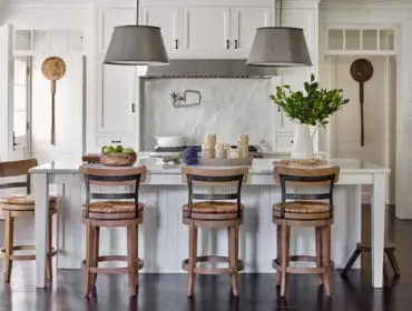 White kitchen with marble backsplash, white island with three wood and woven bar stools, and pendant lights.