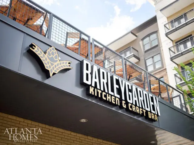 Barleygarden Kitchen & Craft Bar sign with a hop and barley logo, viewed from below with a building and outdoor seating area in the background.