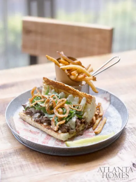 Steak sandwich with arugula, melted cheese, and crispy onion straws, served with french fries and a pickle spear on a rustic wooden table.