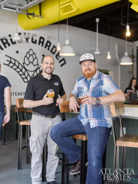 Two men stand and sit at a bar in a brewery with a "BarleyGarden" logo on the wall behind them. The man on the left wears a black polo shirt and khaki pants, holding a beer. The man on the right wears a blue plaid shirt and jeans, also holding a beer.