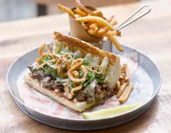 A steak sandwich with melted cheese, crispy onions, and arugula served on a metal plate with a side of french fries in a metal holder and a pickle spear.