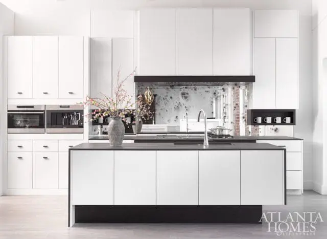 Modern white kitchen with a dark island, stainless steel appliances, and a distressed mirrored backsplash. A vase with pink blossoms sits on the island.