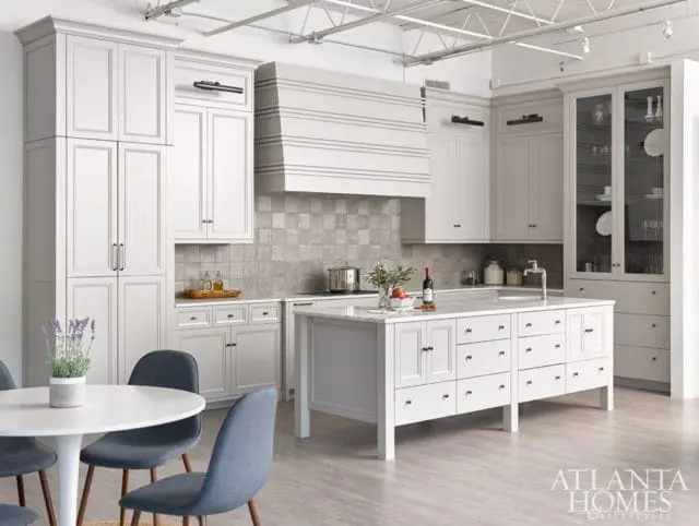 White kitchen with custom cabinetry, marble countertops, a large island with drawers, and a tiled backsplash. A dining table with chairs is in the foreground.