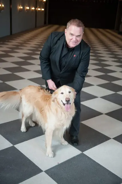 Man in a black suit and black shirt with a red lapel pin standing with a golden retriever dog on a black and white checkered floor.