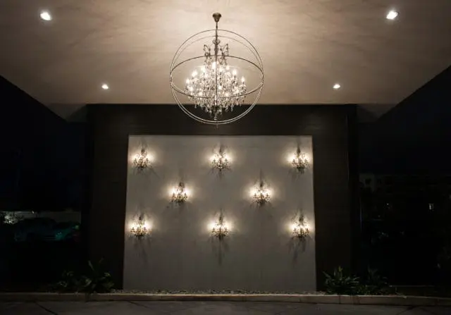 Modern porte cochere with a large, circular metal-framed chandelier hanging above a wall featuring eight ornate sconces casting dramatic shadows.