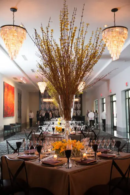 Elegant dining setup in a prefunction room with tables set with tan linens, dark glassware, and tall floral arrangements of yellow flowers and branches. Crystal chandeliers hang from the ceiling.