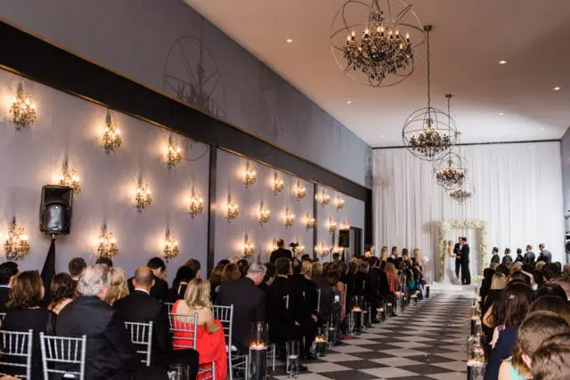 Wedding ceremony with guests seated on chairs arranged in rows on a black and white checkered floor, facing a floral arch where the bride and groom stand. Ornate chandeliers hang from the ceiling, and wall sconces with crystal accents line the side wall.