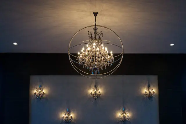 A grand chandelier with multiple lights and crystal accents hangs from the ceiling within a large circular metal frame. Below, five smaller wall sconces with similar crystal details illuminate a textured wall.