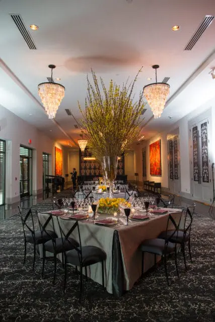 Formal dining room with a long table set for an event, featuring a large floral arrangement, chandeliers, and dark patterned carpet.