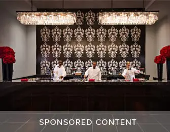 Three servers stand behind a dark granite bar with a black and white damask patterned wall behind them. Two large crystal chandeliers hang overhead. Red floral arrangements are on either side of the bar.