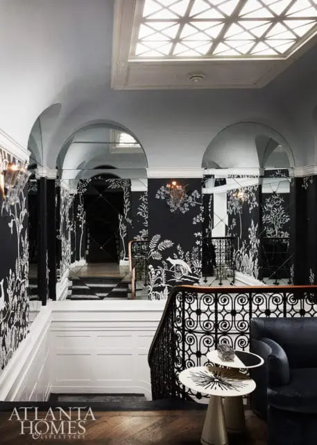 Hallway with black and white botanical wallpaper, mirrored arches, a skylight, and a staircase with ornate railing.