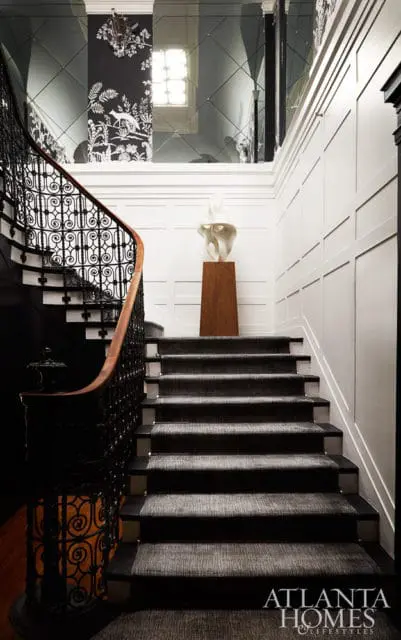 Elegant staircase with black wrought iron railing, wood handrail, and dark patterned carpet runner. White paneled walls and black floral wallpaper accent the space, with a modern sculpture atop a wooden pedestal.