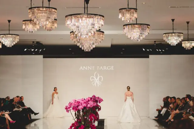 Wedding dress runway show with models in white gowns, audience seated on the sides, and ornate crystal chandeliers overhead.