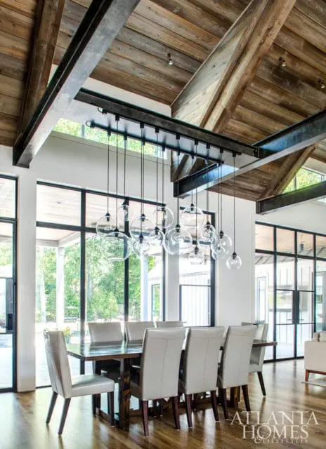 Dining room with a large wooden table and upholstered chairs, featuring a vaulted ceiling with exposed wooden beams and a cluster of glass pendant lights. Large windows offer views of greenery.
