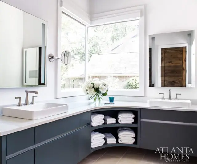 Modern bathroom with a curved dark gray vanity, white countertops, vessel sinks, and a large window overlooking trees. Shelves hold neatly folded white towels.