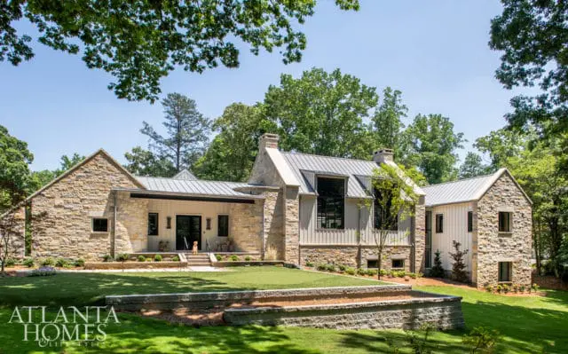 Modern farmhouse with stone and vertical siding, metal roof, and large windows, set in a lush green yard with mature trees.