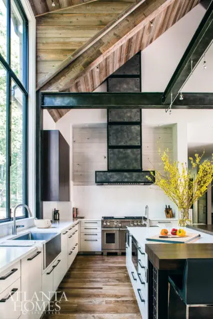 Modern kitchen with vaulted wood-paneled ceiling, exposed steel beams, stainless steel range, white cabinetry, and a large farmhouse sink.