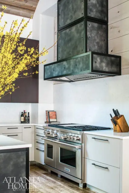 Modern kitchen with stainless steel Wolf range, custom dark metal range hood, white quartz countertops, and light gray cabinetry. A vase of yellow forsythia branches is on the left.
