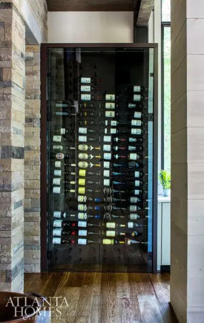 Glass-front wine cellar with rows of wine bottles, flanked by stacked stone and wood paneling.