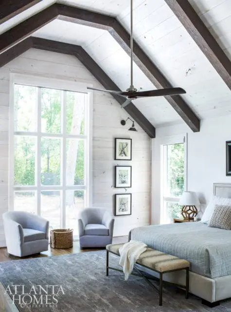 Bedroom with vaulted ceiling featuring exposed dark wood beams and white shiplap. Large windows overlook trees. Includes a king-size bed, two gray armchairs, a bench, and artwork of deer.