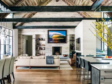 Great room with vaulted wood-paneled ceiling, exposed steel beams, stone fireplace with wave artwork, sectional sofa, dining table, and kitchen island with bar stools.